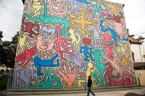 Un mur immense, un peintre bonhomme couleur, des silhouettes vives dansent sur fond blanc. Une femme passe devant cette fresque joyeuse.