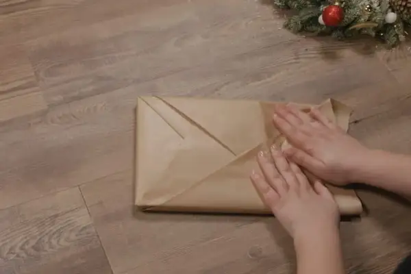 Emballage cadeau livre en papier kraft, prêt pour Noël. Les mains ajustent les plis pour une finition parfaite.