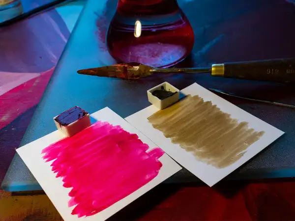 Des petits pots de peinture acrylique, un pourpre intense et un brun terreux, prêts à mélanger pigments. Une spatule et un flacon de liquide rouge complètent la scène.
