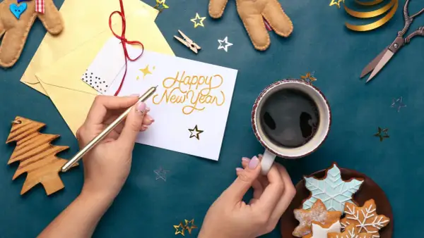 Mains écrivant "Happy New Year" sur une carte, avec une tasse de café et des biscuits.
