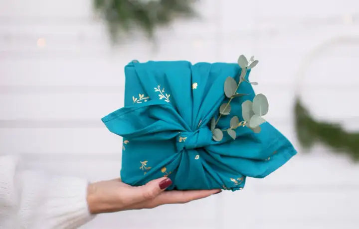 Préparer Noël : un cadeau joliment emballé dans un tissu bleu canard orné de motifs dorés, décoré d'une branche d'eucalyptus.